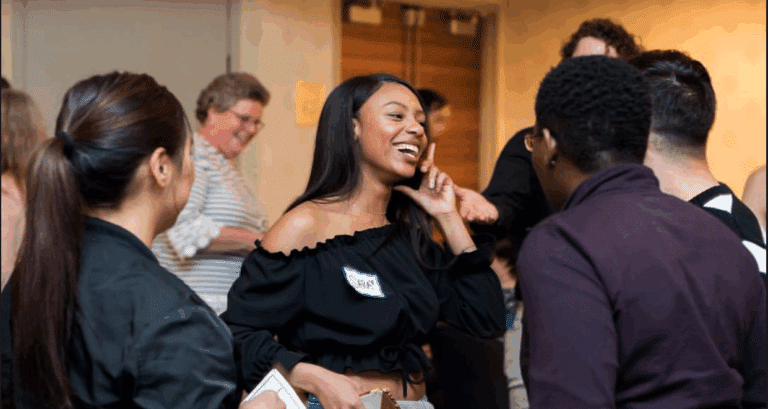 A group of people socialize indoors. A woman in a black off-the-shoulder top is smiling and talking to others, holding a notebook. The atmosphere appears casual and friendly.