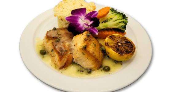 A plated meal featuring chicken breast with a light sauce and capers, a serving of rice, steamed broccoli and carrots, a grilled lemon half, and a purple edible flower garnish.
