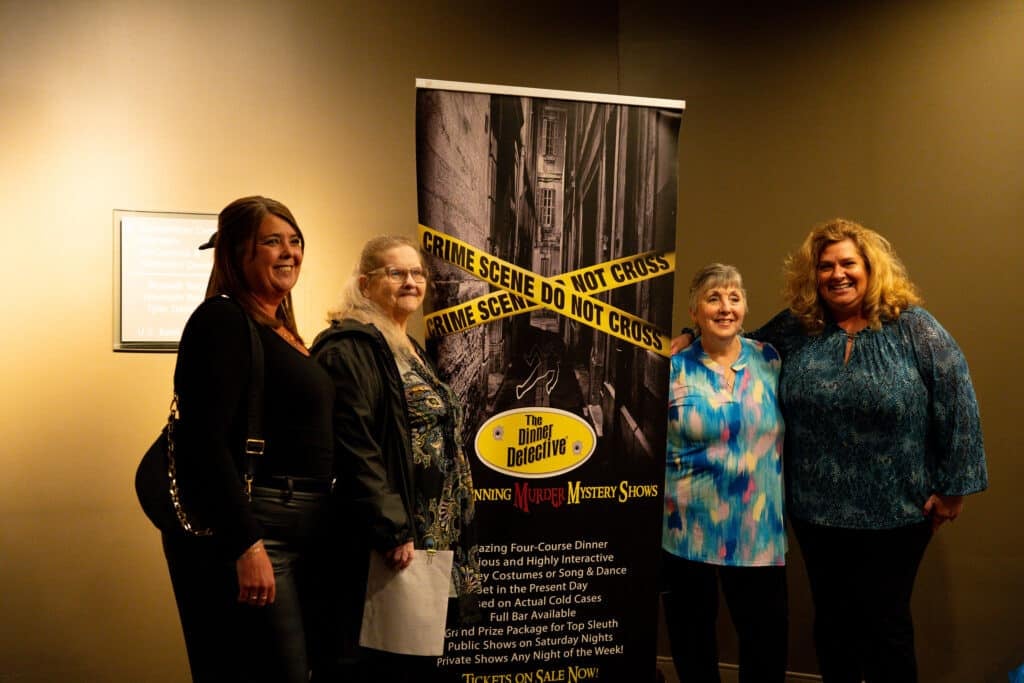 Four women smiling and posing next to a The Dinner Detective Murder Mystery Show banner at an Uptown Charlotte venue. One guest is holding a clue sheet, representing the interactive "hidden actor" experience in the Queen City.