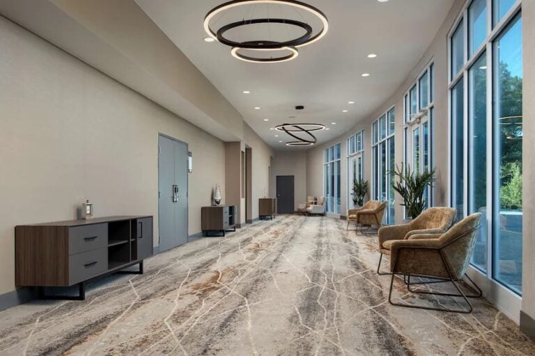 A modern hallway with large windows, beige walls, patterned carpet, brown chairs, potted plants, wooden cabinets, and circular ceiling lights. Natural light fills the space, creating a welcoming atmosphere.