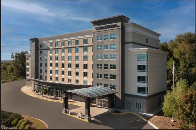 A modern, multi-story hotel building with large windows and a covered entrance sits surrounded by trees and landscaping under a clear sky. The sign on the building reads DOUBLETREE BY HILTON.