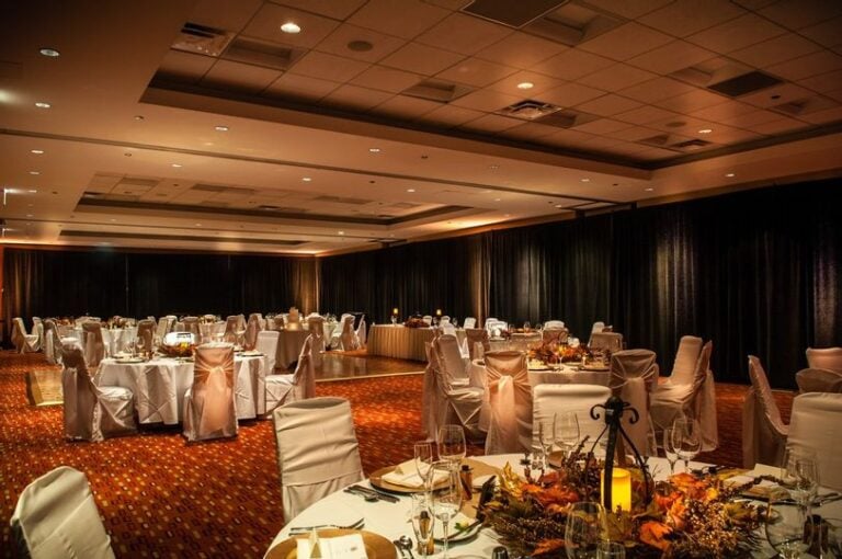 A large banquet hall set for an event, with round tables covered in white cloths and chairs draped in white covers. Warm lighting, autumn-themed centerpieces, and a long head table at the back. The room is empty of people.