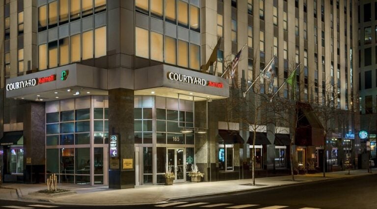 The image shows the exterior of a Courtyard by Marriott hotel at night. The building is well-lit, with large glass windows, multiple flags above the entrance, and streetlights illuminating the sidewalk.