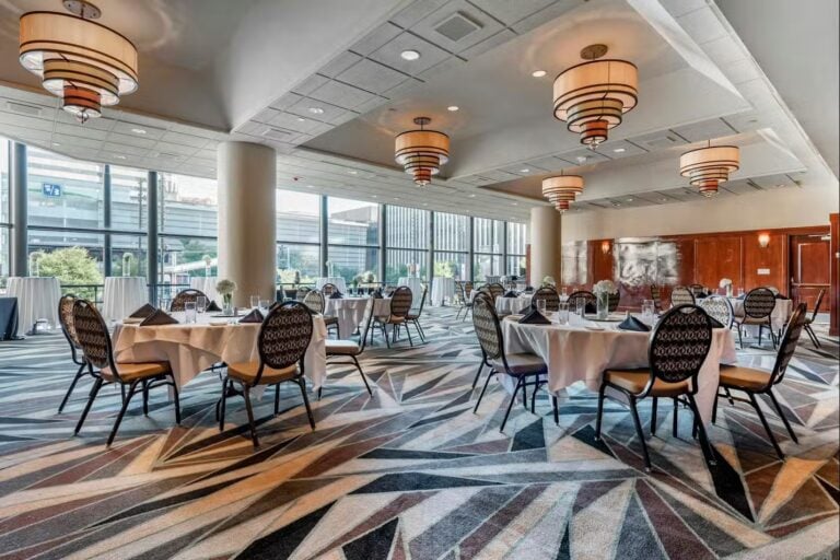 A spacious banquet hall with large windows, round tables covered with white tablecloths, cushioned chairs, geometric-patterned carpet, and modern hanging light fixtures. Some tables are set near windows overlooking an urban scene.