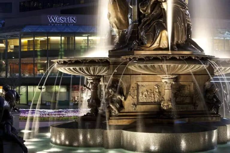 A bronze fountain with cascading water streams is illuminated at night in front of a glass-fronted Westin hotel building. Detailed reliefs and statues adorn the fountain’s base.