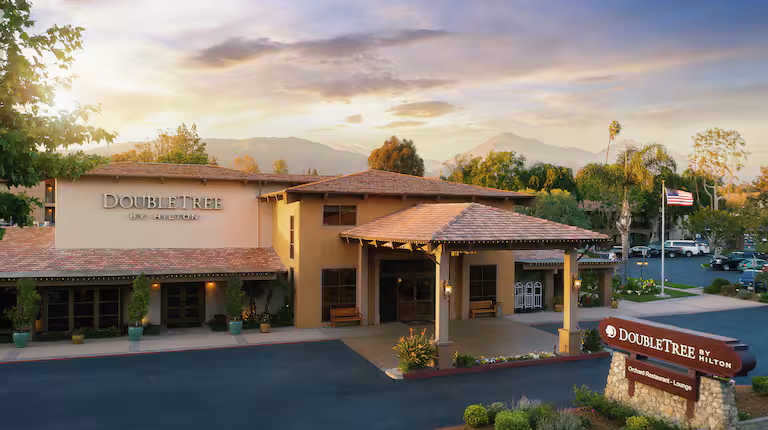 Exterior view of DoubleTree by Hilton hotel with a welcoming entrance, red-roofed building, front sign, greenery, and mountains in the background at sunset.