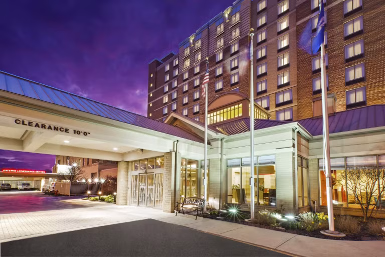 Hotel entrance at dusk with well-lit windows, a canopy displaying a 10-foot clearance sign, flagpoles, and a driveway. The sky is a deep purple, and the building appears modern and welcoming.