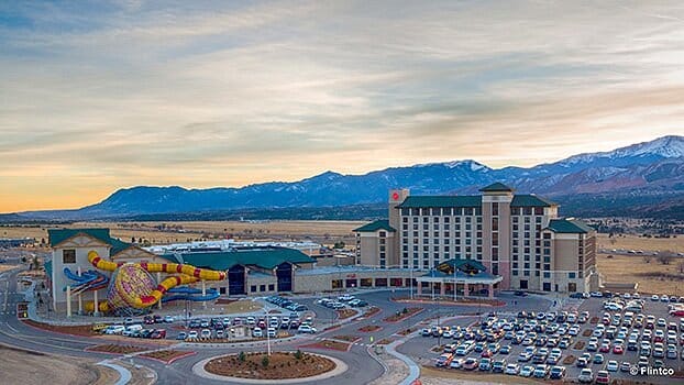 Aerial view of a large hotel and casino complex with a sprawling parking lot, featuring a colorful water slide, set against a backdrop of open plains and distant mountains under a cloudy sky.