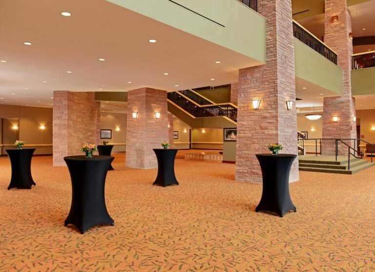 Spacious hotel lobby or event area with brick columns, black cocktail tables with small floral arrangements, patterned carpet, and a staircase leading to an upper level. Warm lighting creates an inviting atmosphere.