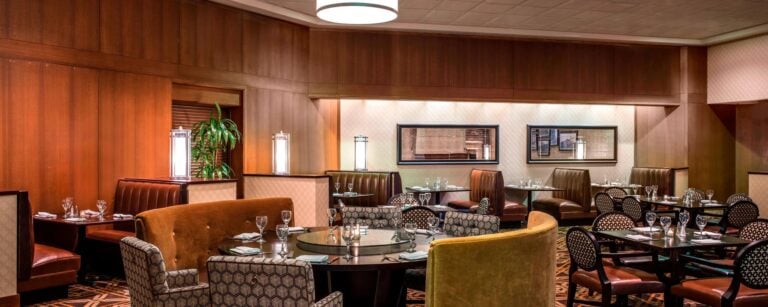 A cozy restaurant interior with wood-paneled walls, round and rectangular tables set for dining, patterned chairs, brown leather booths, soft lighting, and decorative mirrors on the walls.