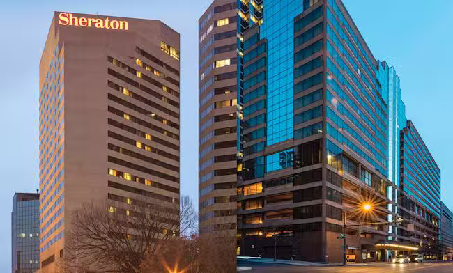 Two modern high-rise buildings at dusk, one with the Sheraton sign at the top. Both buildings have many windows, reflecting the evening sky, and are surrounded by city streets and some trees.