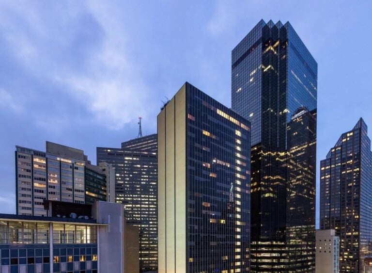 A cityscape at dusk showing several modern office buildings with glass windows, some lights on inside. The sky is cloudy and blue, and the tallest building reflects the evening light.