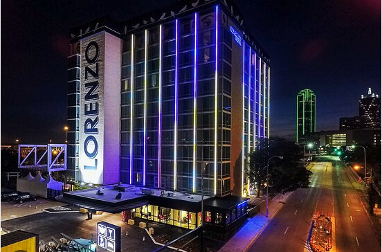 A tall building with “LORENZO” written vertically on its side, illuminated by colorful lights at night. The surrounding city street is quiet, and skyscrapers with green lights are visible in the background.