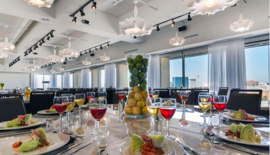 Elegant banquet table set with plates of salad, assorted wine glasses, and a tall glass vase filled with lemons and limes. Floor-to-ceiling windows show a cityscape in the background. White pendant lights hang overhead.