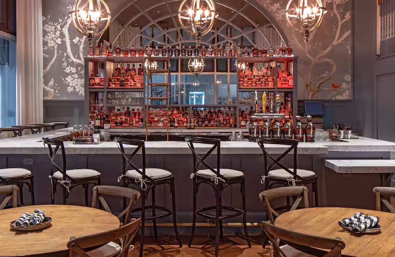 Elegant restaurant bar with high chairs, marble countertop, round wooden tables, napkins, and shelves stocked with bottles. Warm lighting and decorative wall art create an inviting, sophisticated atmosphere.