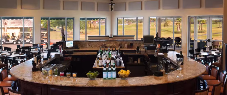 Spacious bar area with a marble countertop, surrounded by chairs, bottles of drinks, and bowls of fruit. Large windows offer views of a golf course and outdoor seating in the background.