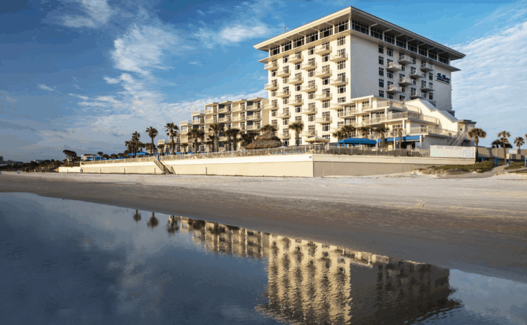 A modern beachfront hotel with balconies overlooks a sandy beach and is reflected in shallow water under a blue sky with scattered clouds. Palm trees line the property.