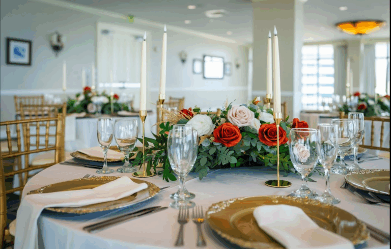 A round table set for a formal event with gold chairs, gold-rimmed plates, white napkins, crystal glasses, and a centerpiece of red and white roses with greenery and tall, unlit taper candles.