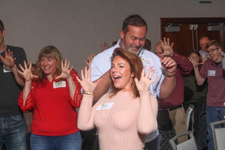 A group of adults standing in a room, smiling and raising their hands with fingers spread wide, appearing to participate in an interactive activity or exercise.