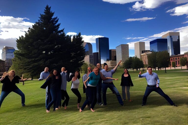 A group of people pose playfully on a grassy field in front of tall trees and modern city buildings under a partly cloudy sky.