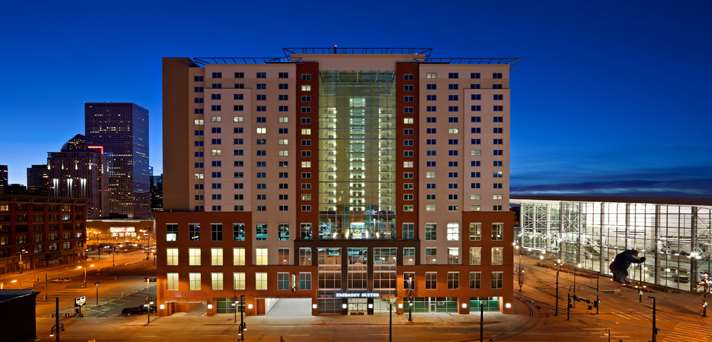 A tall, modern building with many lit windows stands in a cityscape at dusk, next to a large glass-walled structure. The sky is deep blue and streetlights illuminate the surrounding area.