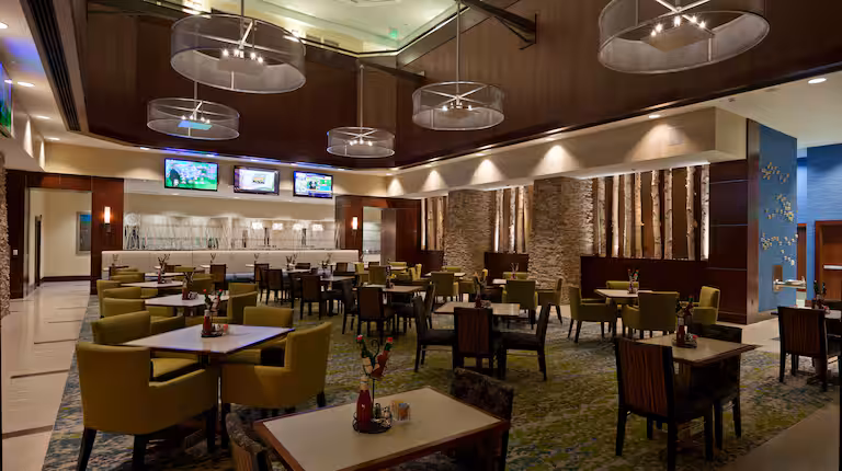Modern hotel dining area with green chairs and square tables, contemporary chandeliers, stone accent walls, and several TV screens above a white counter. The space has a warm, inviting atmosphere with artistic decor.