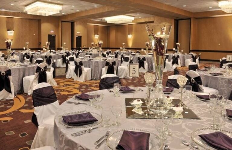 A large banquet hall set for an event with round tables covered in white cloths, each with purple napkins, glassware, and elegant centerpieces. Chairs have white covers with purple sashes, and table number 5 is visible.
