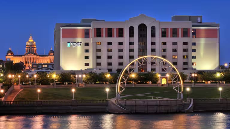A modern Embassy Suites hotel building is illuminated at dusk, with a riverside park and decorative lights in the foreground. A domed, historic government building is visible in the background.