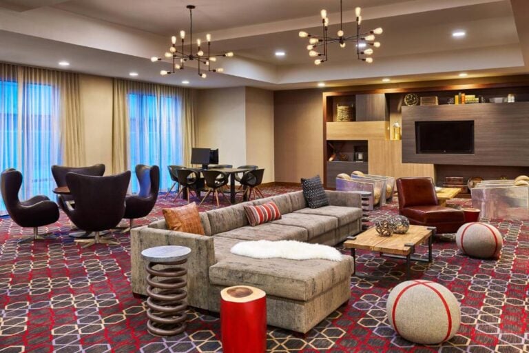 Modern lounge with patterned carpet, contemporary sofas, round chairs, playful poufs, and stylish lighting fixtures. A TV is mounted on a built-in wall unit with shelves, and two computers sit by large windows with sheer curtains.