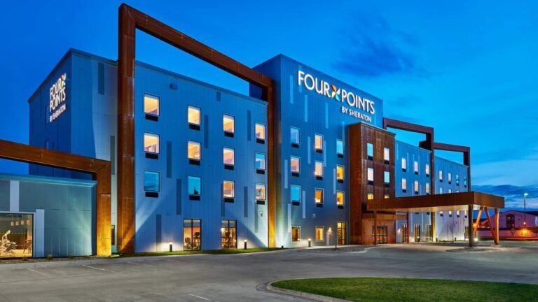 Modern Four Points by Sheraton hotel building at dusk with illuminated windows and signage, featuring unique brown architectural frames and a covered entrance, set against a blue evening sky.