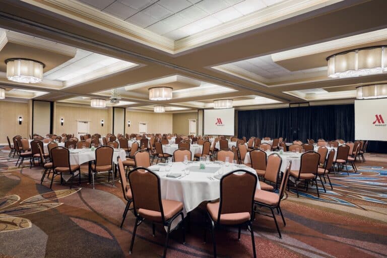 A large, empty conference room with round tables covered in white tablecloths and surrounded by chairs, set up for an event. The room features patterned carpet, chandeliers, and Marriott logos on two projector screens.