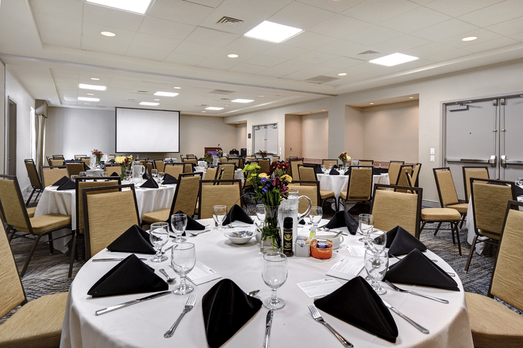 A banquet hall set up for an event with round tables covered in white tablecloths, black napkins, and floral centerpieces. Each table has glasses, menus, and tableware. Background features a projection screen and empty chairs.