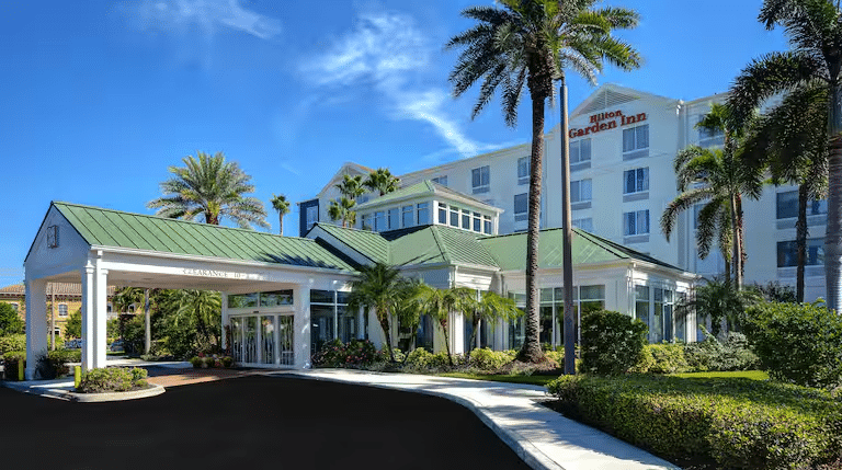 Exterior of a Hilton Garden Inn with a green roof, surrounded by palm trees under a clear blue sky. The building is white with several windows and a welcoming entrance. Landscaping features include neatly trimmed bushes and a paved driveway.