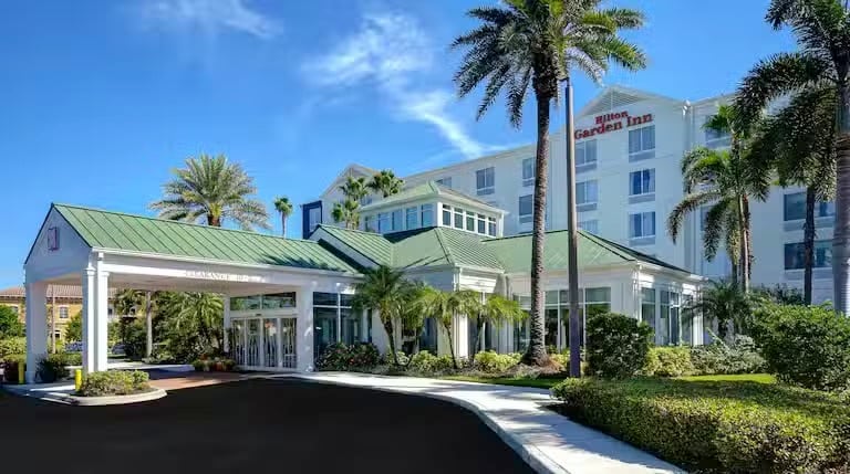 The exterior of a Hilton Garden Inn hotel, featuring a green-roofed entrance surrounded by lush palm trees and shrubs under a bright blue sky.
