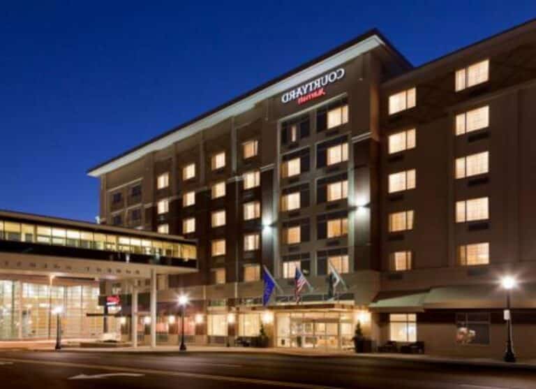 Courtyard by Marriott hotel exterior at night, with lights glowing from windows, flags at the entrance, and a skybridge connecting to another building. The street in front is empty.