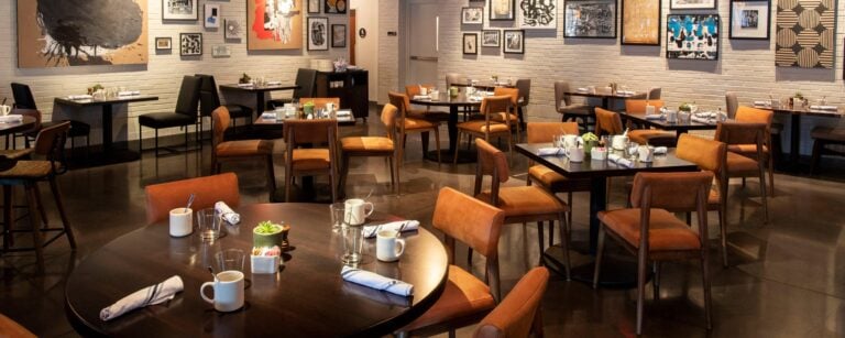 A modern restaurant dining area with empty tables and chairs, set with mugs, napkins, and small decorations. Artworks and photographs decorate the white brick walls, creating a cozy and inviting atmosphere.
