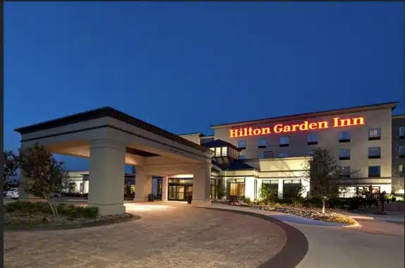 A Hilton Garden Inn hotel is shown at dusk, with illuminated signage on the building. The entrance features a covered driveway, landscaped areas, and a well-lit, welcoming atmosphere.