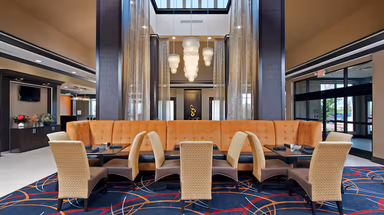 Modern hotel lobby with high ceilings, hanging light fixtures, a long orange cushioned booth, beige patterned chairs, glass tables, and a colorful carpet with circular patterns. Large windows let in natural light.