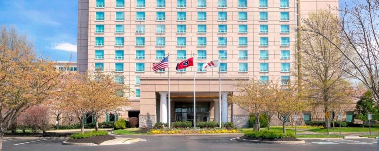 A multi-story hotel with rows of windows, a landscaped entrance, and five flagpoles displaying the U.S., Tennessee, and other flags. Flower beds and small trees line the front driveway.