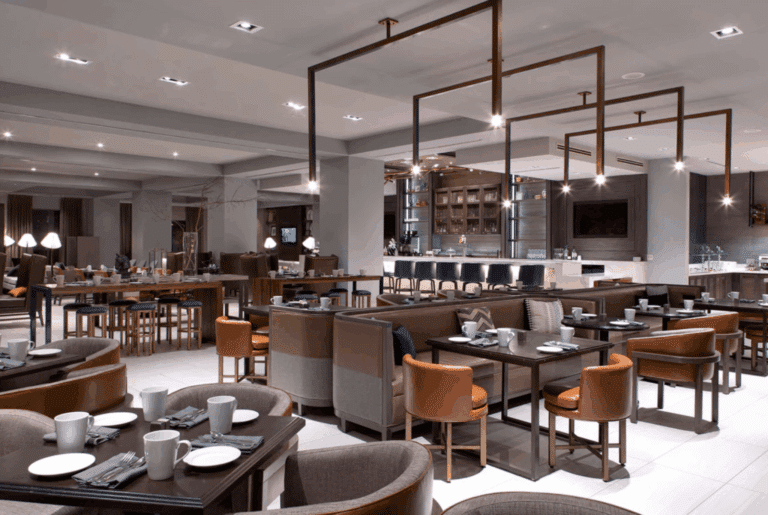 Modern restaurant interior with neatly arranged tables and chairs, neutral color tones, minimalist decor, overhead lighting, and a bar area in the background. The space appears empty, with no people present.