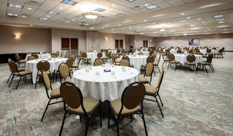 A large banquet hall with round tables covered in white tablecloths, each surrounded by brown cushioned chairs. The room is well-lit with ceiling lights and chandeliers, and the carpet has a light, patterned design.