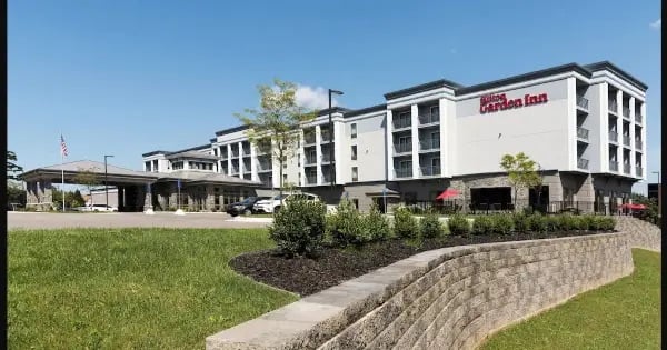 A modern Hilton Garden Inn hotel with multiple stories, large windows, an entrance canopy, and landscaped grounds with green grass and shrubs under a clear blue sky.