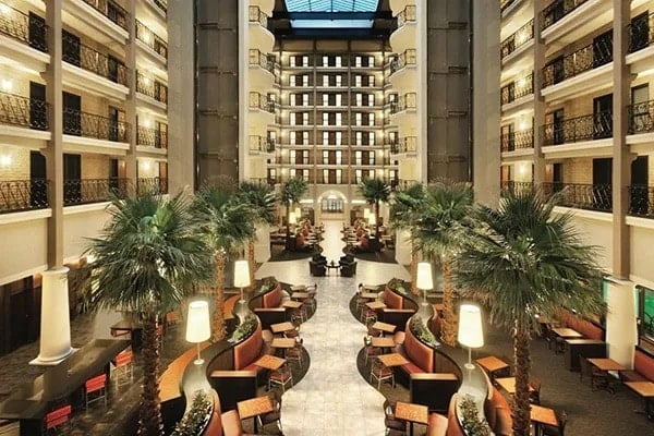 Spacious hotel atrium with multiple floors, balconies, palm trees, modern seating areas, and warm lighting under a glass ceiling, creating an inviting and luxurious atmosphere.