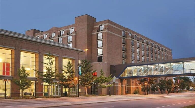 A large brick hotel building with multiple floors stands behind a glass skywalk that connects to a modern structure with large windows, set along a quiet city street at dusk.