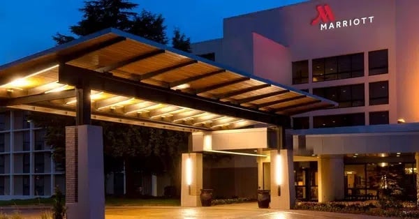 A well-lit entrance canopy leads to a modern Marriott hotel building at dusk, with the red Marriott logo illuminated on the facade. Trees and windows are visible in the background.