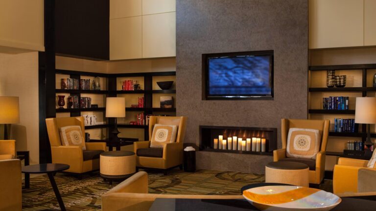 A cozy lounge area with armchairs, bookshelves, a wall-mounted TV, and decorative candles in a modern fireplace. Warm lighting and contemporary decor create a comfortable, inviting atmosphere.
