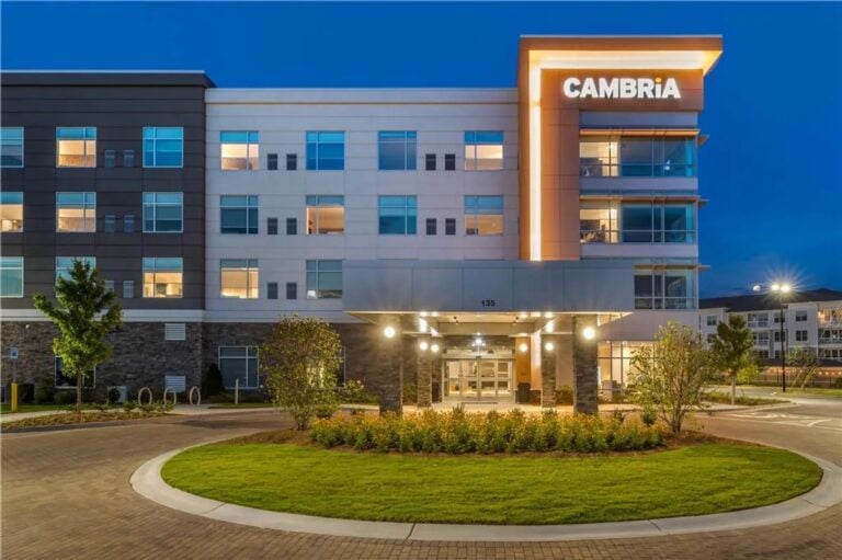 Modern hotel building at dusk with illuminated CAMBRiA sign, large windows showing lit rooms, main entrance under a covered drive, landscaped greenery, and a circular driveway in the foreground.
