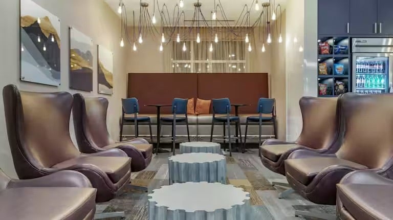 Modern lounge area with brown leather chairs, abstract coffee tables, blue cushioned chairs at a high table, wall art, hanging pendant lights, and a snack vending area with drinks in a cooler on the right.