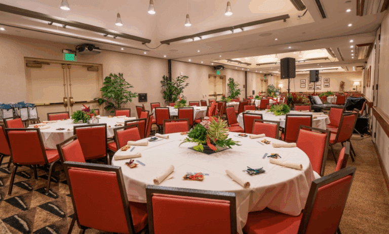 A banquet hall set up with round tables covered in white tablecloths, each surrounded by red chairs and decorated with green centerpieces. Rolled napkins and small gifts are placed at each seat.
