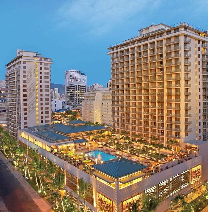 Aerial view of a brightly lit resort hotel at dusk, featuring a central outdoor swimming pool surrounded by palm trees, with tall buildings and a cityscape in the background.